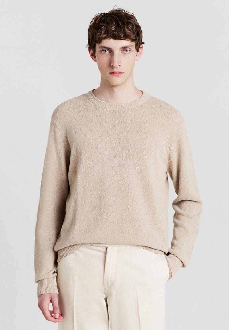 Junger Mann mit kurzen, lockigen braunen Haaren, der einen beigen Strickpullover und crèmefarbene Hosen trägt und mit einer Hand in der Tasche vor einem weißen Hintergrund steht.