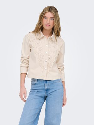 Junge Frau mit welligem blondem Haar, die eine beige Jacke mit Knöpfen und Stickerei mit Ösen sowie hellblaue Jeans trägt, steht vor einem weißen Hintergrund.