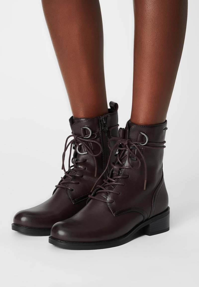 Bottines en cuir marron foncé avec lacets, fermeture éclair latérale et petit talon bloc portées sur des jambes à peau foncée sur fond blanc.