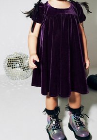 Vestido de terciopelo morado con mangas cortas y lazos en los laterales. Botas brillantes y con iridiscencias, con cordones, volantes y acentos en negro.