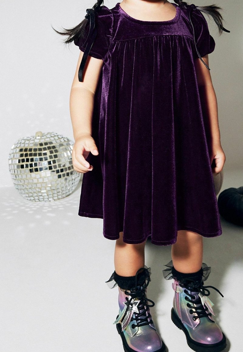 Vestido de terciopelo morado con mangas cortas y lazos en los laterales. Botas brillantes y con iridiscencias, con cordones, volantes y acentos en negro.