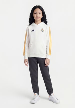 Jeune personne aux longs cheveux foncés portant un sweat à capuche blanc du Real Madrid avec des rayures orange et un pantalon noir, debout sur un fond blanc.