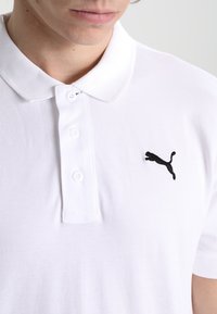 Bílá polo košile vyrobená z texturované tkaniny s knoflíkovou légou na tři knoflíky a černým vyšitým logem Puma na levé hrudi.