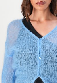 Lichtblauwe, doorschijnende cardigan met een losse brei-textuur, met een V-hals, knopen aan de voorkant en een donkere onderlaag zichtbaar.