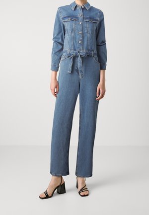 Femme portant une combinaison bleu denim à manches longues, avec des boutons sur le devant, une ceinture à nouer à la taille, et des sandales à talon bloc noires à bout ouvert sur un fond uni.