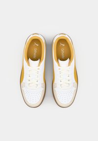 Hvite sneakers med gule detaljer og en tan gummisåle. Teksturert skinnoverdel med perforeringer, snøring og Ortholite-merket inni.