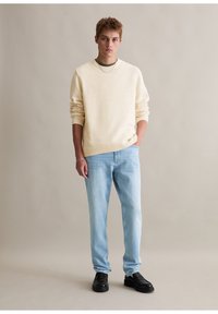 Helle beige strukturierte Sweatshirt, entspannte Passform, Rundhalsausschnitt. Kombiniert mit hellblauen Jeans, schmale Passform, und schwarzen Schuhen. Einfaches Design mit neutralen Farben.