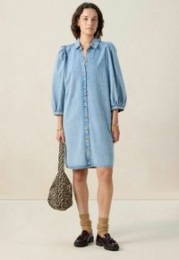 Femme portant une robe en denim arrivant au genou avec des manches bouffantes, tenant un sac à imprimé léopard, assortie à des chaussettes marron et des mocassins foncés.