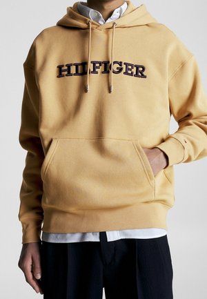 Beżowa bluza z kapturem z czarnym haftem "HILFIGER", noszona na białej koszuli, z jedną ręką w przedniej kieszoni i widocznymi czarnymi spodniami.