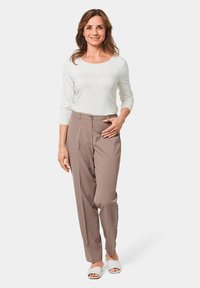 Beige op maat gemaakte broeken met een gladde textuur, gecombineerd met een witte top met lange mouwen. Het model staat in een minimalistische omgeving, met witte slide sandalen.