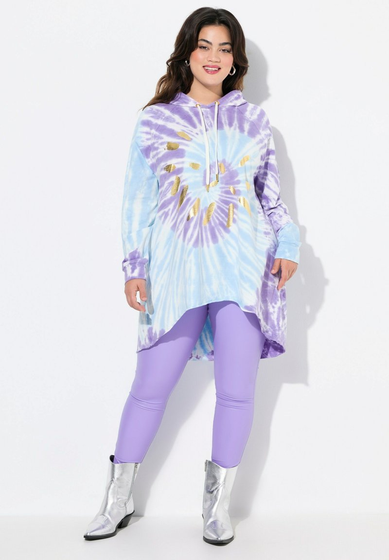 T-shirt met tie-dye in blauw en paars met gouden accenten; lange mouwen, asymmetrische zoom. In combinatie met paarse legging en zilveren laarzen.