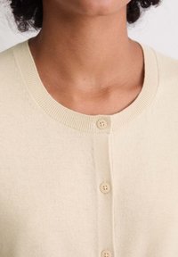 Cremefarbene gestrickte Strickjacke mit rundem Ausschnitt und zwei strukturierten Knöpfen auf der Vorderseite, mit gerippten Kanten und weichem Stoff.