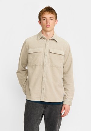 Jeune homme portant une chemise beige en velours côtelé boutonnée avec deux poches poitrine et un pantalon gris foncé, debout sur fond blanc.