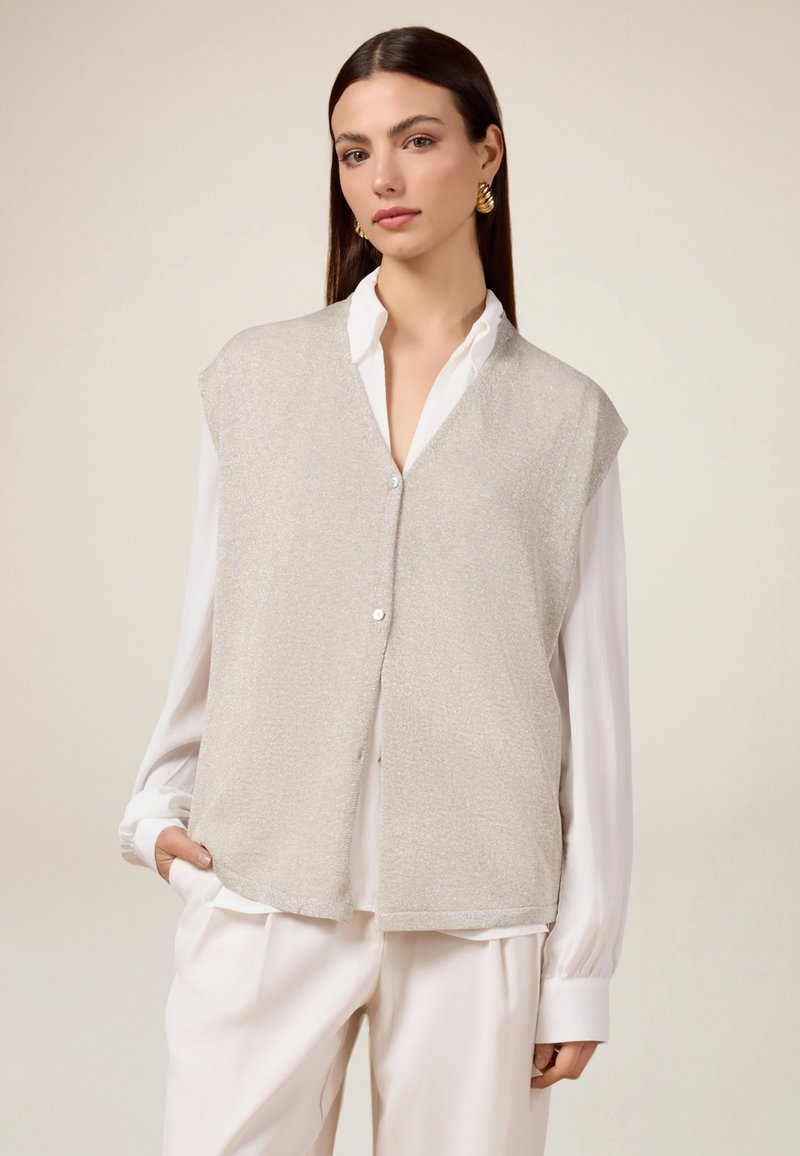 Giubbetto senza maniche grigio chiaro in un tessuto testurizzato, con chiusura a bottoni, indossato sopra una camicia bianca, abbinato a pantaloni chiari.