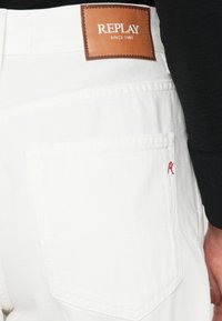 Τα λευκά παντελόνια από denim διαθέτουν καφέ δερμάτινο λογότυπο με την επιγραφή "REPLAY" στη μέση. Ένα κόκκινο κεντητό διακοσμητικό είναι ορατό στην πίσω τσέπη.