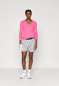 Suéter rosa com nervuras em V, calções xadrez preto e branco, sapatos brancos e uma luva de golfe. Design minimalista, tecido leve.