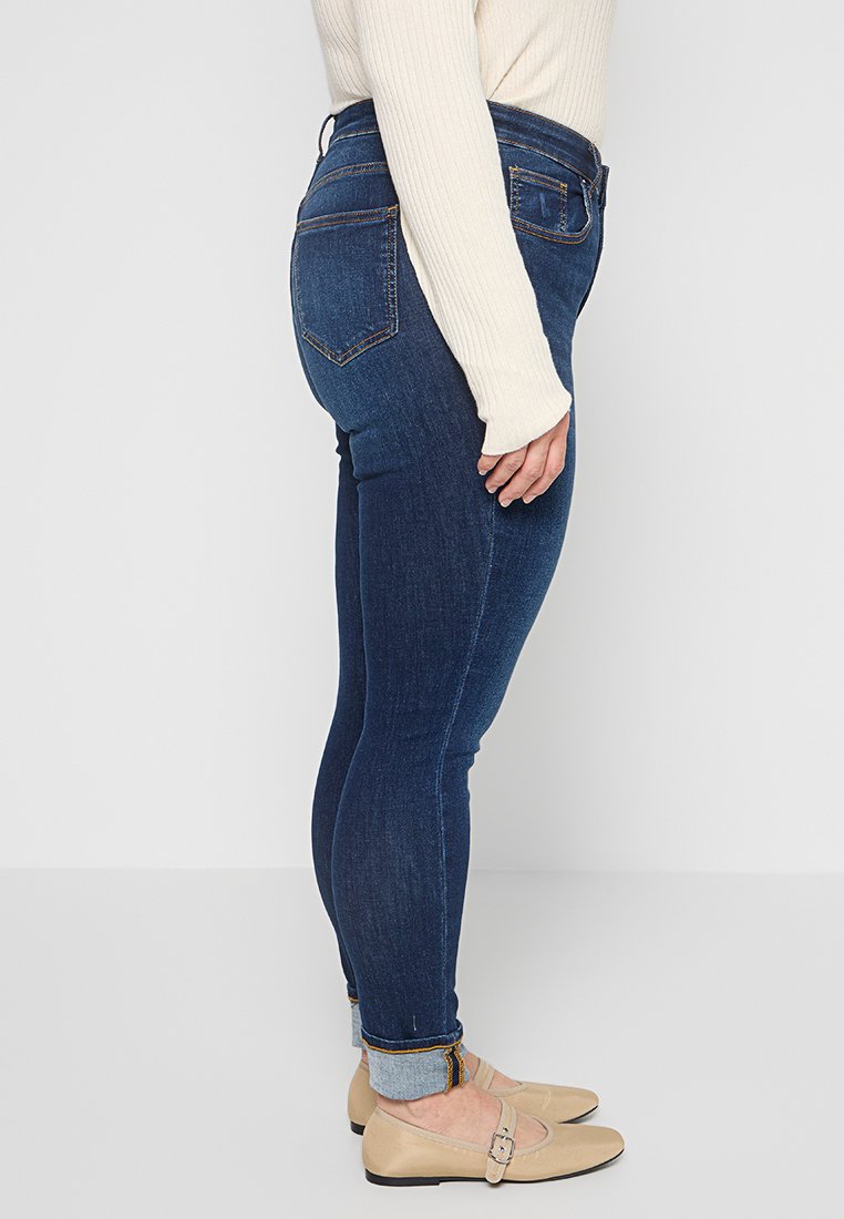 Dunkelblaue Skinny-Jeans mit mittelhohem Bund, die eine glatte Denim-Oberfläche und dezente Gesäßtaschen aufweisen, mit aufgerollten Säumen und Stickereien.
