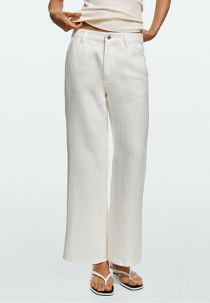 Person trägt lockere weiße Hose und weiße Sandalen und hebt mit einer Hand ein beiges Oberteil leicht an, um die Taille zu zeigen.