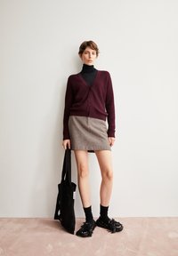 Bordeaux cardigan met knopen, zwarte coltrui, grijze tweed rok, zwarte handtas en zwarte schoenen met franjes op een lichtroze vloer.