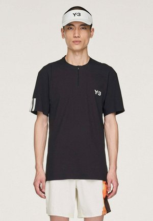 Mann trägt ein schwarzes Y-3-Shirt mit Frontreißverschluss, weiße Shorts mit orangefarbenem Detail und ein weißes Y-3-Visier vor einem einfarbigen Hintergrund.