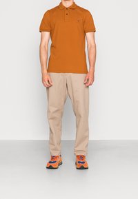 Pomarańczowa koszulka polo z małym logo, beżowe spodnie i kolorowe trampki z niebieskimi i pomarańczowymi akcentami. Outfit jest casualowy i dobrze dopasowany.