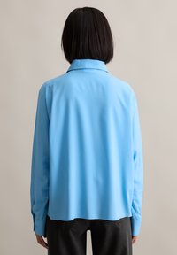 Light blue collared shirt with long sleeves, smooth texture, relaxed fit, and side slits at the hem, paired with dark pants.