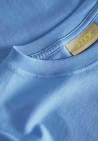 T-shirt in cotone azzurro chiaro con collo a giro. Superficie liscia e bordi ripiegati. Etichetta gialla con il nome del marchio visibile.