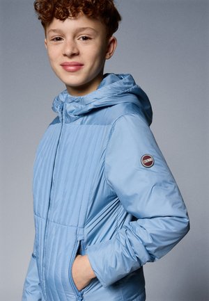 Giacca puffer azzurro chiaro con cappuccio, presenta cuciture verticali, tasche laterali e una patch rotonda con logo sul braccio sinistro. Texture liscia.