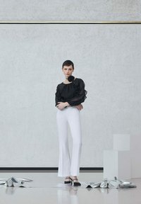 Blusa nera trasparente con dettagli a volant, abbinata a pantaloni bianchi attillati. L'outfit presenta un accento floreale nero e scarpe nere.