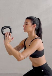 Svart kettlebell holdt med begge hender av en person i svart sports-BH og tights, plassert mot en lys grå bakgrunn.