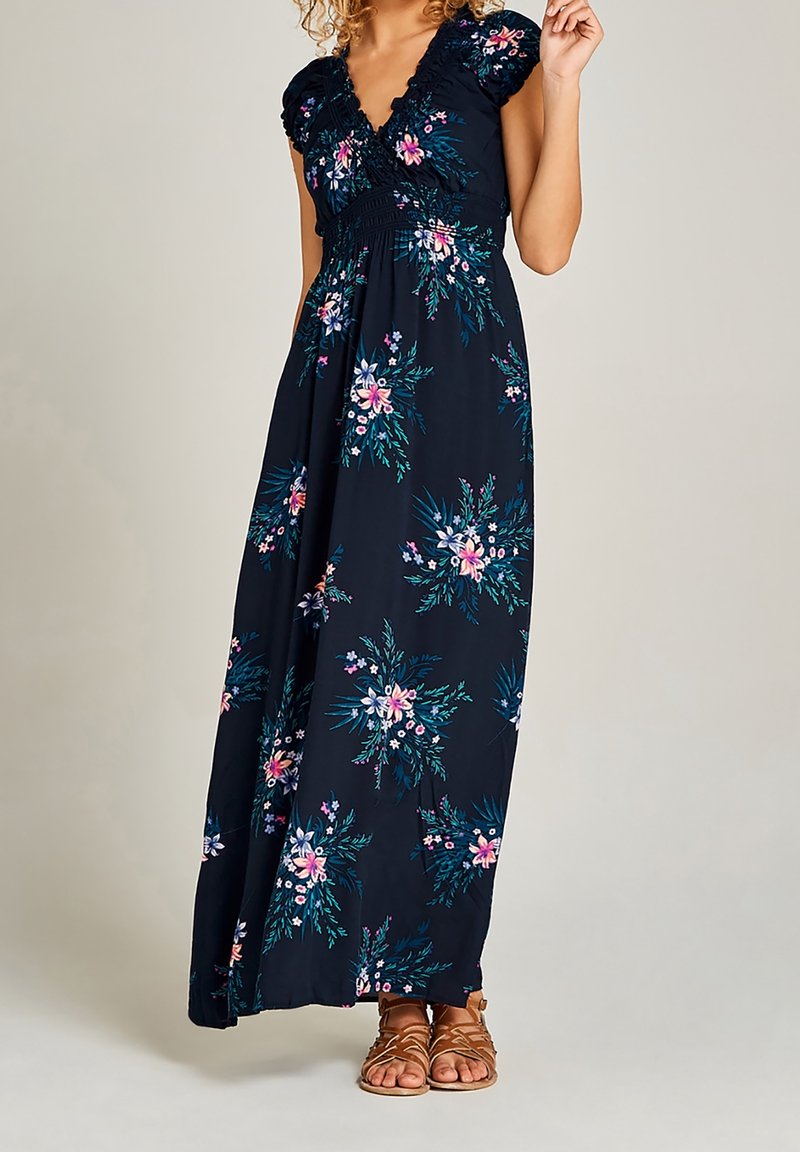 Femme portant une robe maxi fleurie bleu marine avec des manches courtes et des sandales marron à lanières, debout devant un fond uni.