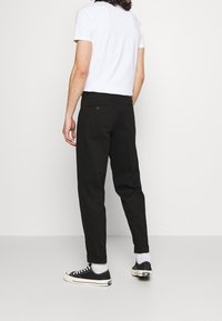 Black trousers with a tapered shape, made from cotton. Features a rolled cuff, two back pockets, and a button closure. Worn with a white t-shirt and sneakers.