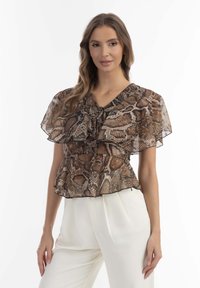 Barna és világosbarna peplum blúz kígyóbőr mintával, áttetsző, fodros ujjakkal, V-nyakkivágással és elől fodros részletekkel. Fehér nadrággal párosítva.