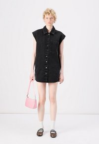 Zwarte denim jurk met kraag, korte mouwen, knopen op de voorkant en een enkele borstzak. Gecombineerd met een roze handtas en schoenen met een patroon.
