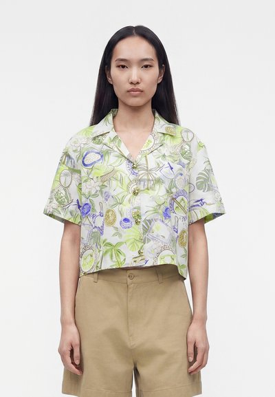 Jeune femme aux longs cheveux foncés portant une chemise à manches courtes à imprimé tropical et un short beige, debout devant un fond uni.