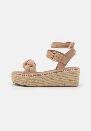 Beige suède sleehaksandaal met gevlochten plateauzool, enkelriem met gesp en geknoopte voorriem.