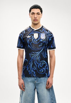 Jeune homme portant un maillot de football argentin noir et bleu avec les logos FIFA et AFA, associé à un jean ample bleu clair, debout sur un fond blanc.