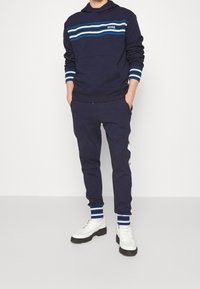 Marinblå hoodie med blå och vita ränder, framficka och matchande marinblå svettbyxor. Vita sneakers med svarta sulor kompletterar outfiten.