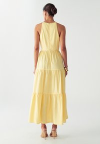 Nainen, jolla on nuttura ja yllään ilman-hihoja oleva keltainen kerroksellinen maxi-mekko, kultaiset renkaat korvissa, käsikoruja ja nudet korkeakorkoiset sandaalit, selkä kohti.