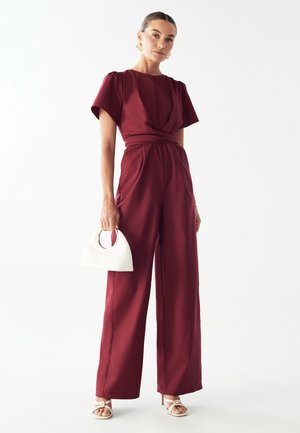 Bordeaux jumpsuit med korte pufærmer, en knyttet talje, bred benudskæring, parret med en hvid handbag og strophæle. Glat tekstur.