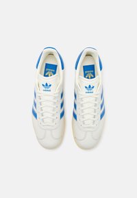 Zapatillas blancas con tres rayas azules, parte superior de cuero y suela de goma. Presenta un logo en la lengüeta y cordones en contraste.