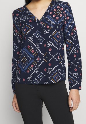 Femme portant un chemisier à manches longues bleu marine avec des motifs géométriques et floraux multicolores, associé à un pantalon noir.