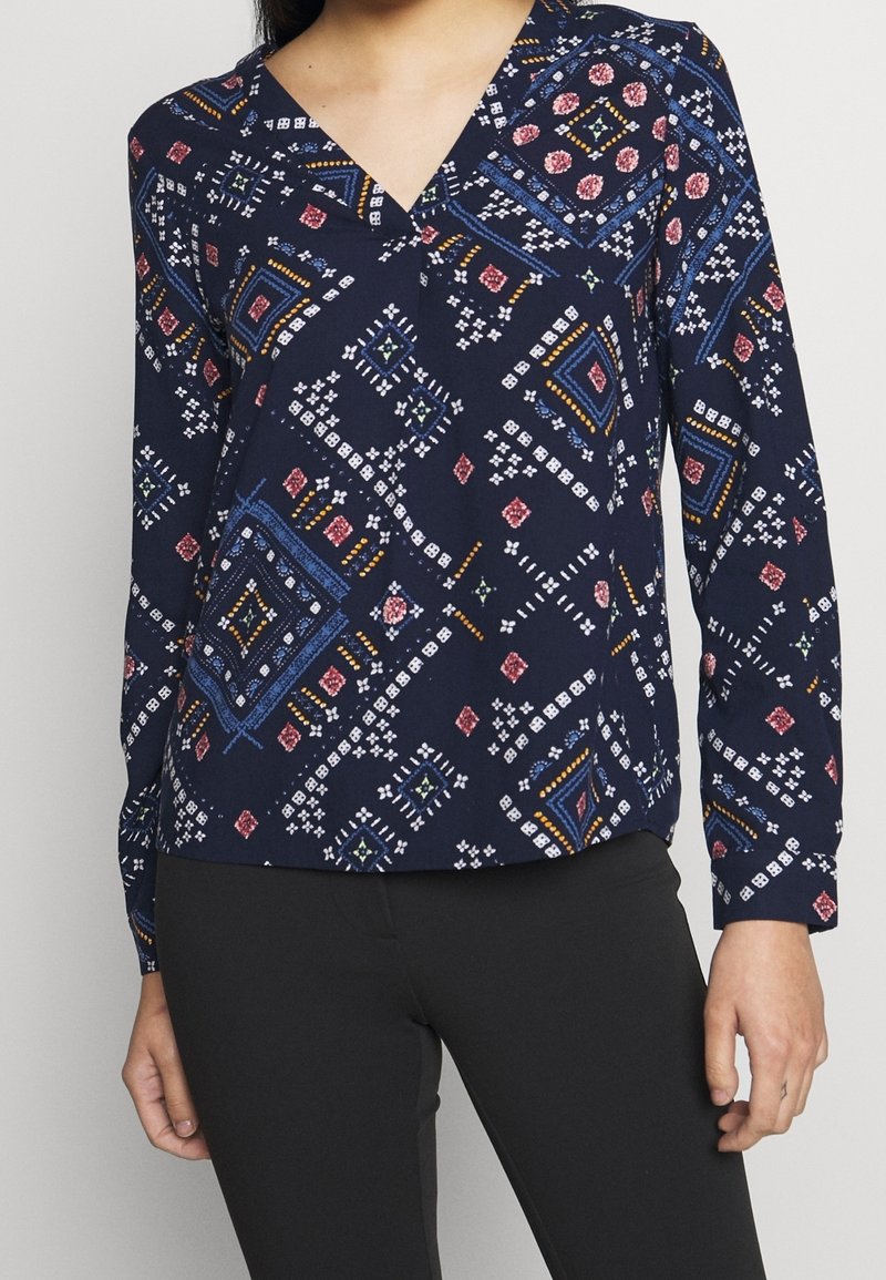 Femme portant un chemisier à manches longues bleu marine avec des motifs géométriques et floraux multicolores, associé à un pantalon noir.
