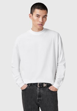 White long-sleeve crewneck shirt made from cotton, paired with dark gray jeans. The shirt features a relaxed fit and ribbed neckline.