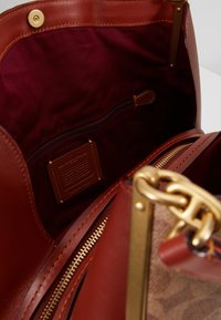 Intérieur du sac à main en cuir en tissu bordeaux profond ; comprend une poche zippée, des ferrures dorées et une étiquette de marque embossée sur le cuir.