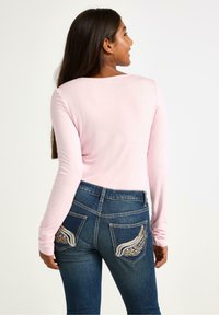 Lichtroze langemouwen top gecombineerd met donkerblauwe jeans met decoratieve stiksels op de achterzakken. Gladde stoftextuur.