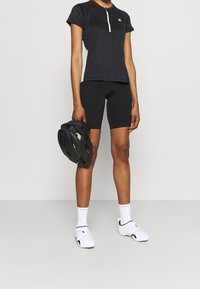 T-shirt de vélo noir à manches courtes avec une fermeture éclair à l'avant, associé à un short de vélo noir. Le modèle tient un casque de vélo noir. Des chaussures de sport blanches sont portées.