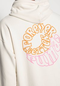 Krémová mikina s kapucňou s veľkým kruhovým grafickým motívom na chrbte, na ktorom je oranžový a ružový text "FOREVER BETTER PUMA". Mäkká látka, voľný strih.