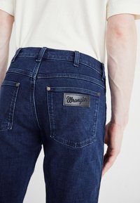 Tamsiai mėlynos džinsinio audinio kelnės su tiesiu kirpimu, su juodu odiniu Wrangler logotipo žymekliu ir metaliniais vinimis ant užpakalinių kišenių.