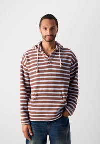 Mann med kort krøllete hår og skjegg iført en brun og lys blå-stripete hettegenser og blå jeans, stående mot en ensfarget bakgrunn.